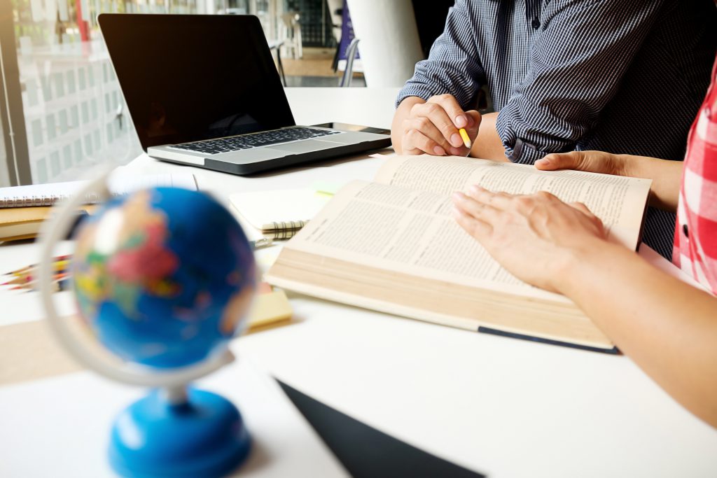 Zbliżenie na ręce dwóch specjalistów pracujących z dużą księgą i laptopem nad projektem obejmującym tłumaczenia techniczne czeski.