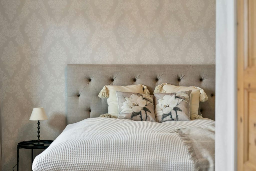 Luksusowe szare łóżko z pikowanym zagłówkiem w sypialni z wzorzystą tapetą, prezentujące jakość, jaką oferuje producent łóżek tapicerowanych.